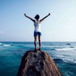A beautiful ocean vista is shown with a woman whose back is to the camera holding her arms up in triumph as she stands at the top of a rock feeling thrilled that ASX 200 shares are reaching multi-year high prices today